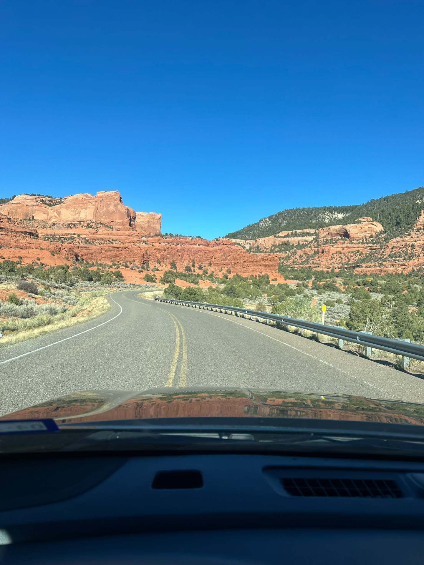A two-lane highway curving through red sandstone cliffs and juniper country under a clear blue sky.