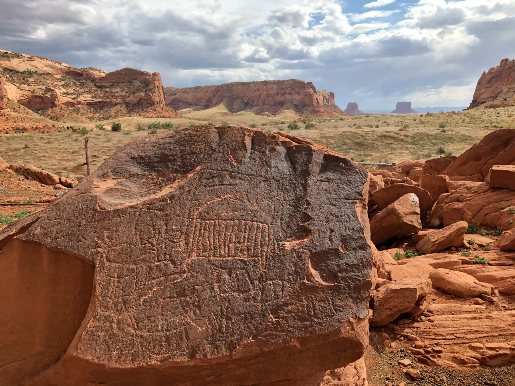 A large sandstone boulder carved with petroglyphs, with red mesas and buttes on the horizon behind it.