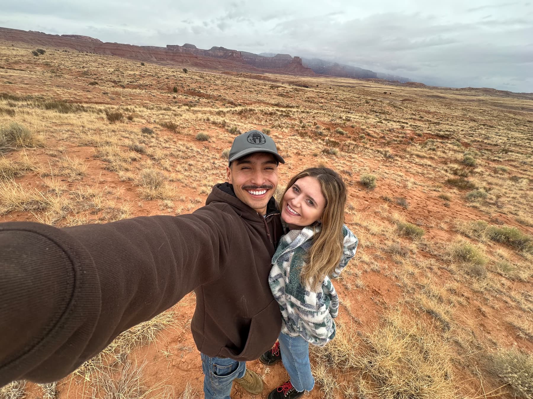Tim and Ann standing together in open high desert grassland with red cliffs and low clouds behind them.