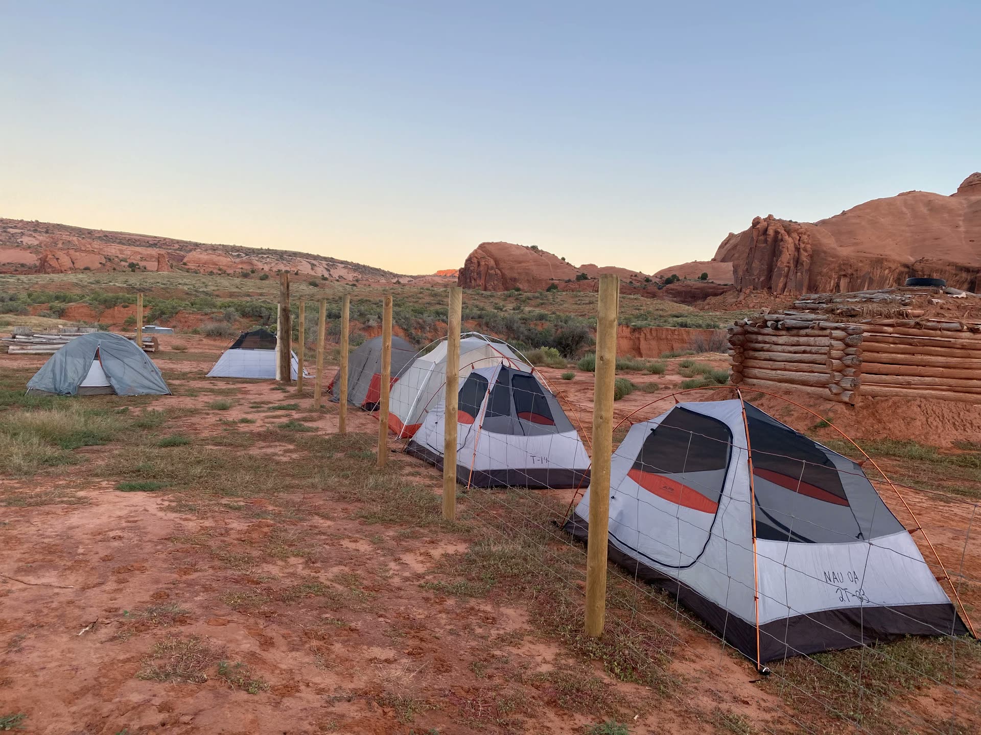 Tents pitched on red sand at first light beside a log hogan, with sandstone bluffs behind.