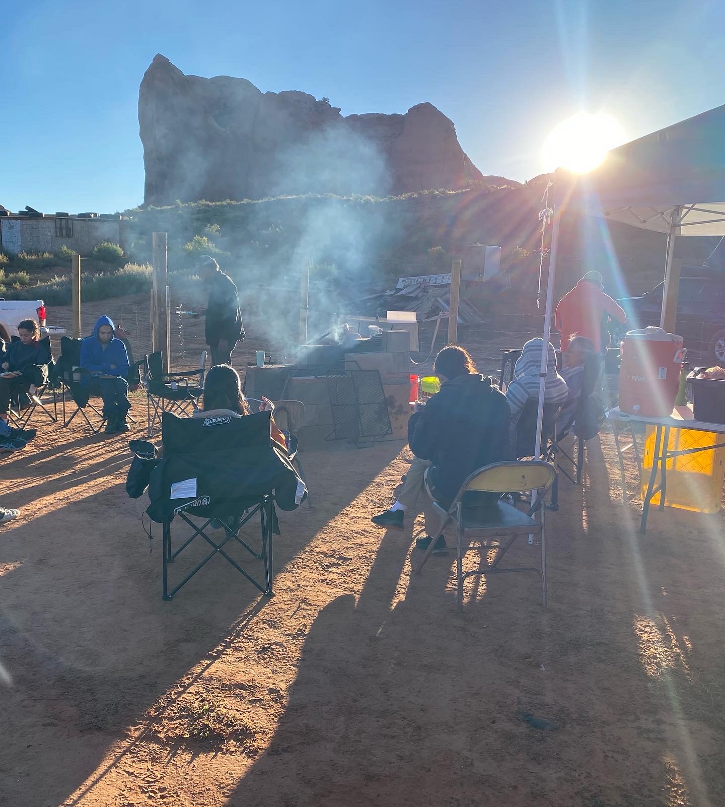 Early morning at camp with woodsmoke rising in front of a red sandstone butte, people seated in camp chairs.