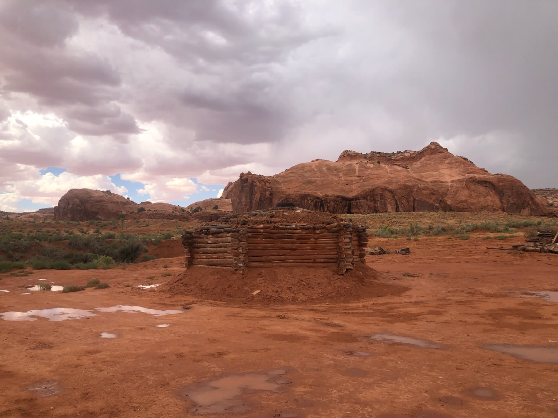 A traditional log-and-earth hogan standing on red sand below sandstone bluffs after a rainstorm.