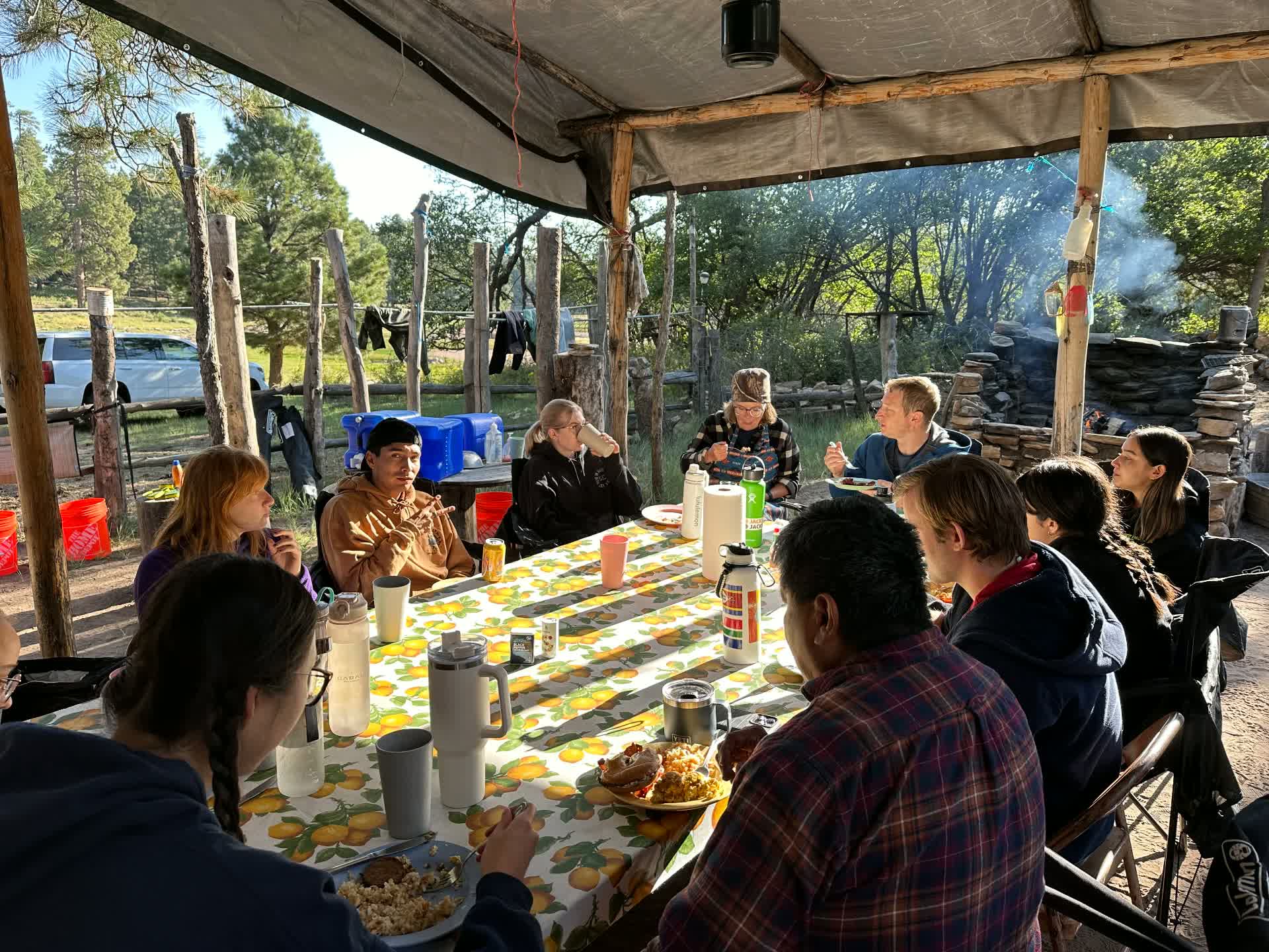 A long table under a shade shelter where a group shares a morning meal outdoors.